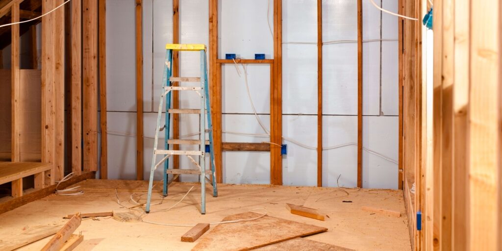 Early stages of a custom home remodeling project showing an unfinished room with wooden framing, insulation between beams, electrical boxes on walls, and a worn step ladder.
