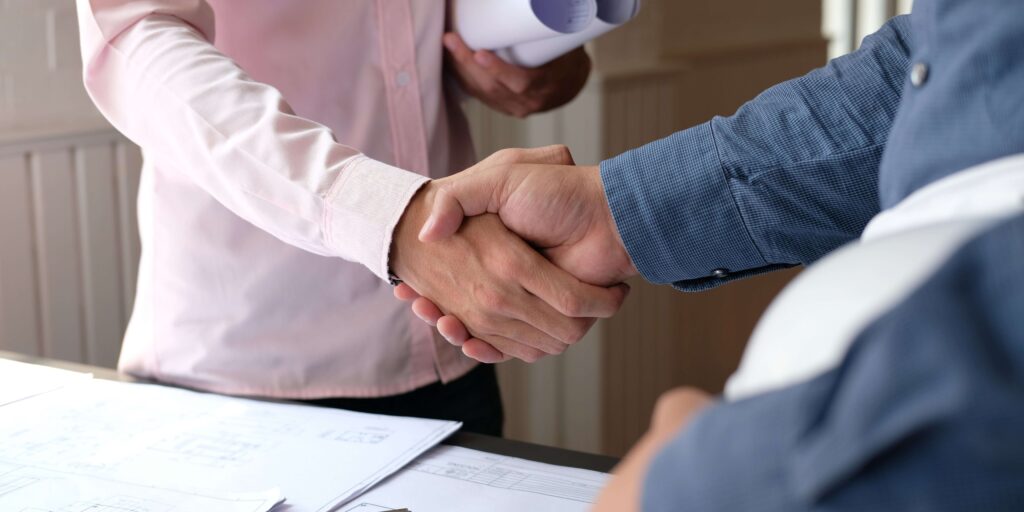 A close-up of a firm handshake between a client and a reliable home remodel contractor, with architectural blueprints and a construction helmet in the background, representing a solid agreement on a new home renovation project.
