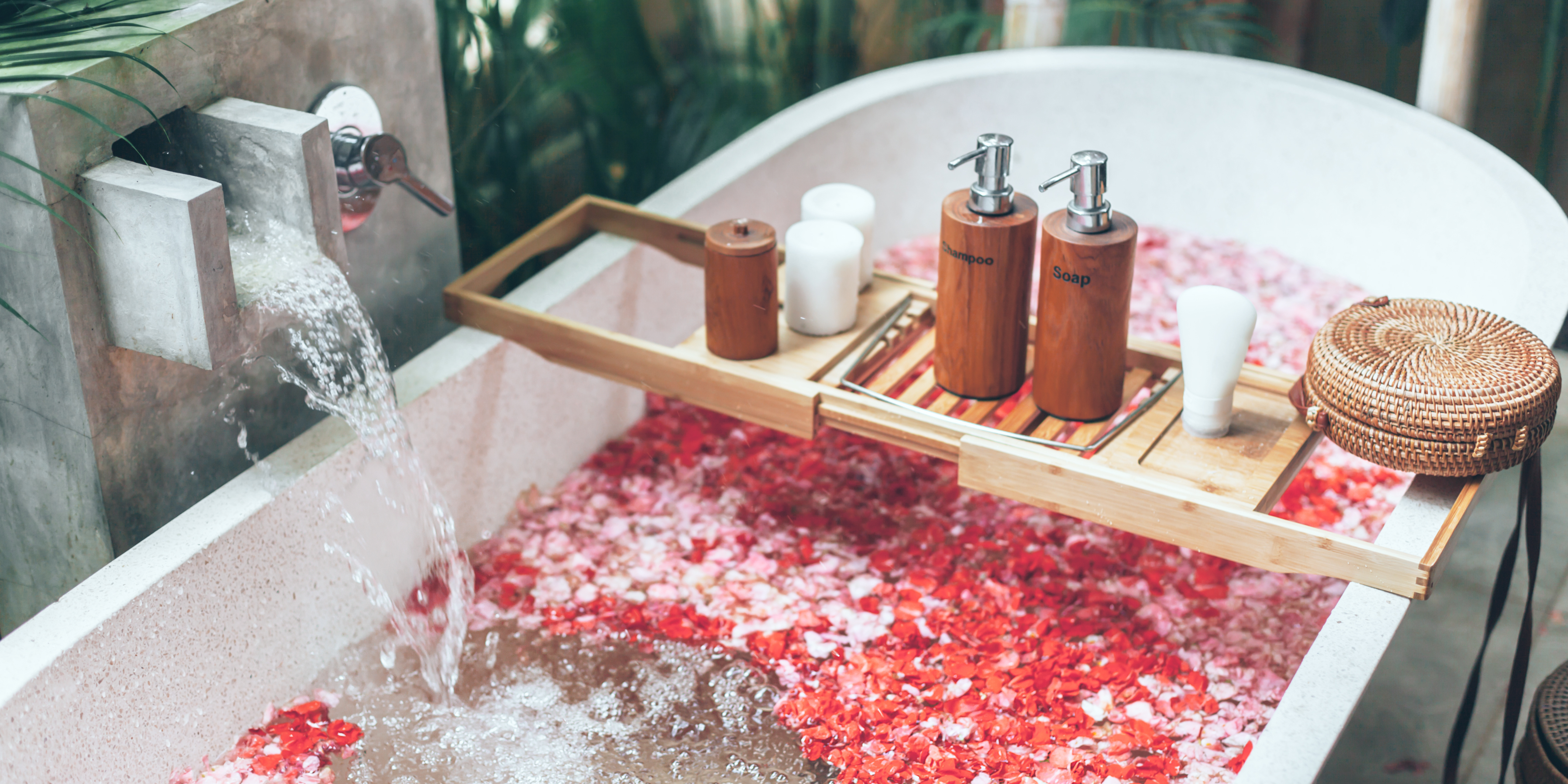 Luxurious full bathroom makeover featuring a curved bathtub with rose petals floating in water, complemented by a wooden bath tray holding eco-friendly bath products, a woven basket, and a serene backdrop of lush greenery.
