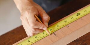 A contractor's hands using a pencil to mark measurements on a wooden board with a yellow tape measure, capturing a detail-oriented task in a complete home remodel project.