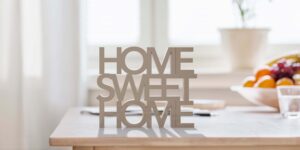 Decorative 'HOME SWEET HOME' sign in a modern kitchen setting with a blurred background of a fruit bowl, symbolizing the comfort and increased home value achieved through a whole house remodel.