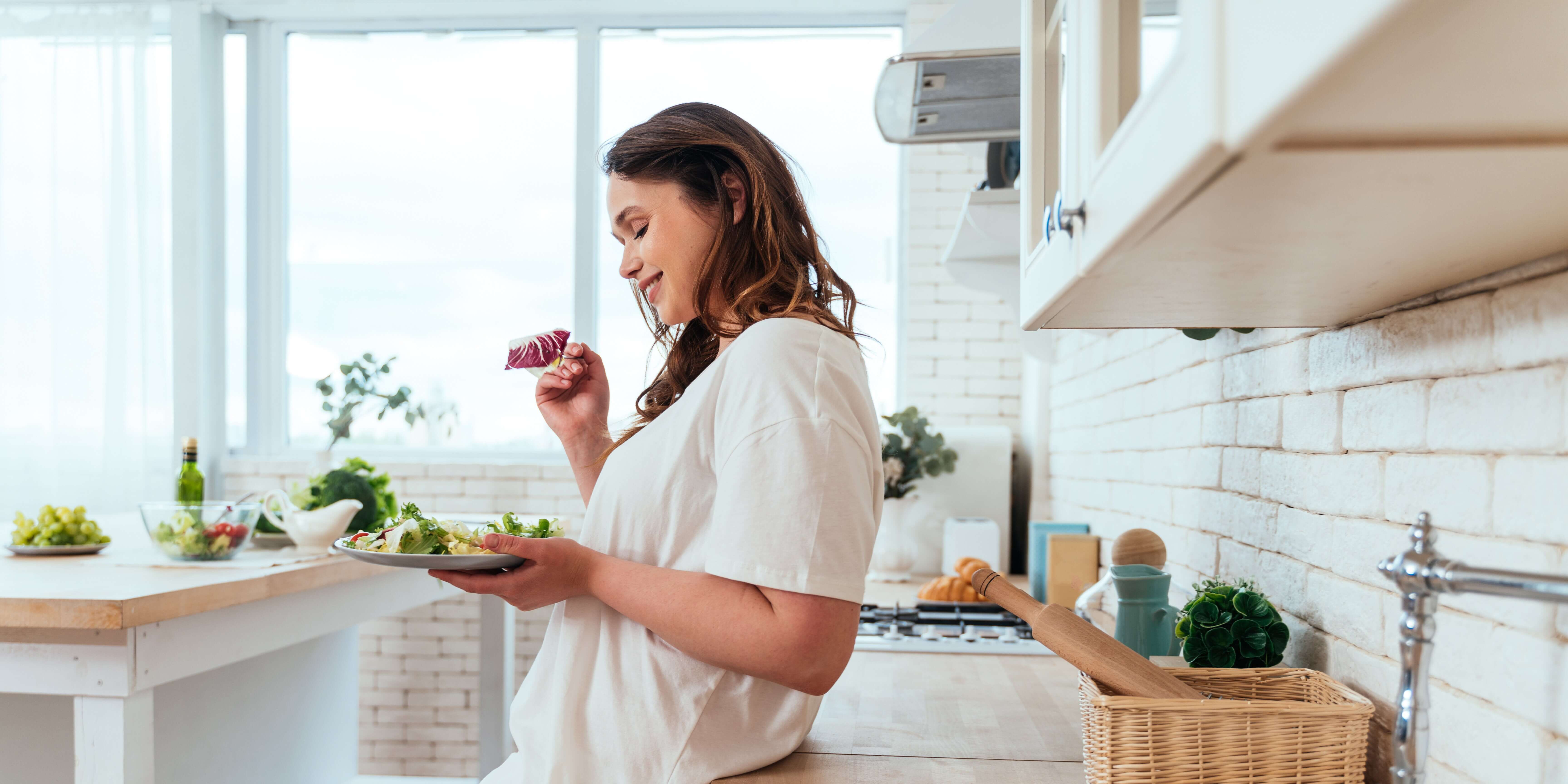 After the custom kitchen remodeling, a woman is admiring a bite of salad in a sunlit kitchen, remodeled with white brick backsplash, sleek wooden countertops, and contemporary appliances, embodying a stylish and functional culinary space.
